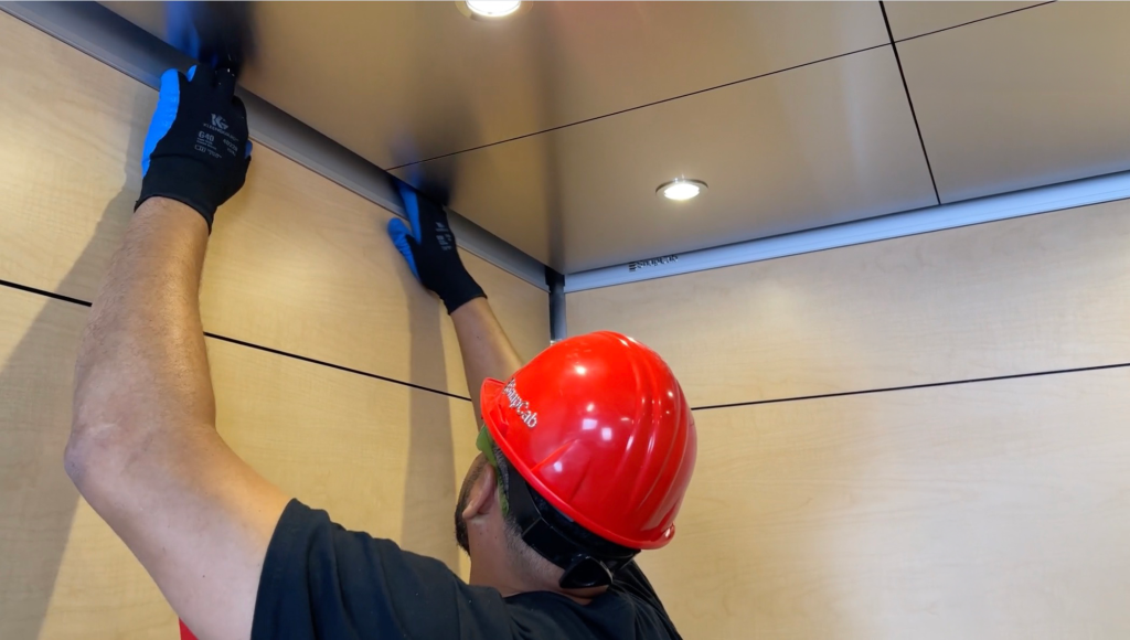 Elevator mechanic wearing a red hard hat and black gloves installs or adjusts metal trim in an elevator interior