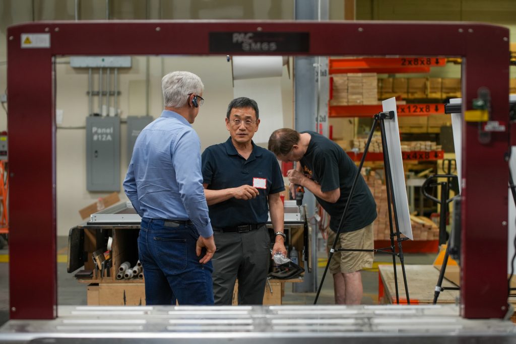 Two men talking in a manufacturing warehouse while a worker builds a panel in the background.