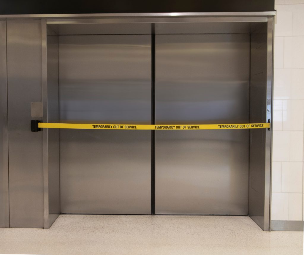 A bright yellow 'Elevator Out of Service' warning banner stretched across modern, brushed stainless steel elevator doors in an office lobby.