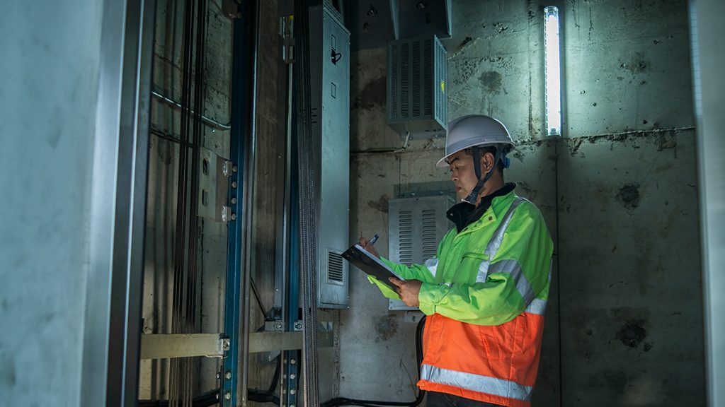 An elevator maintenance technician in a white hard hat and high-visibility safety vest writing on a clipboard during an inspection inside a lift shaft.