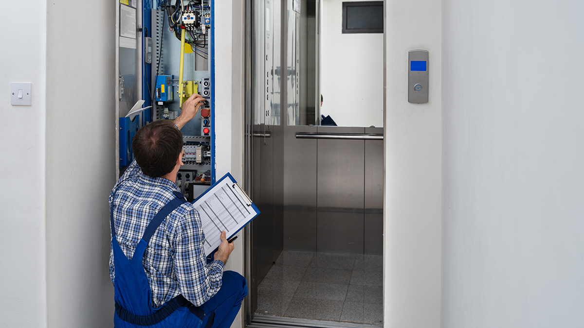 An elevator technician in blue overalls holding a checklist on a clipboard while adjusting wiring inside an open mechanical controller panel next to an open elevator cab