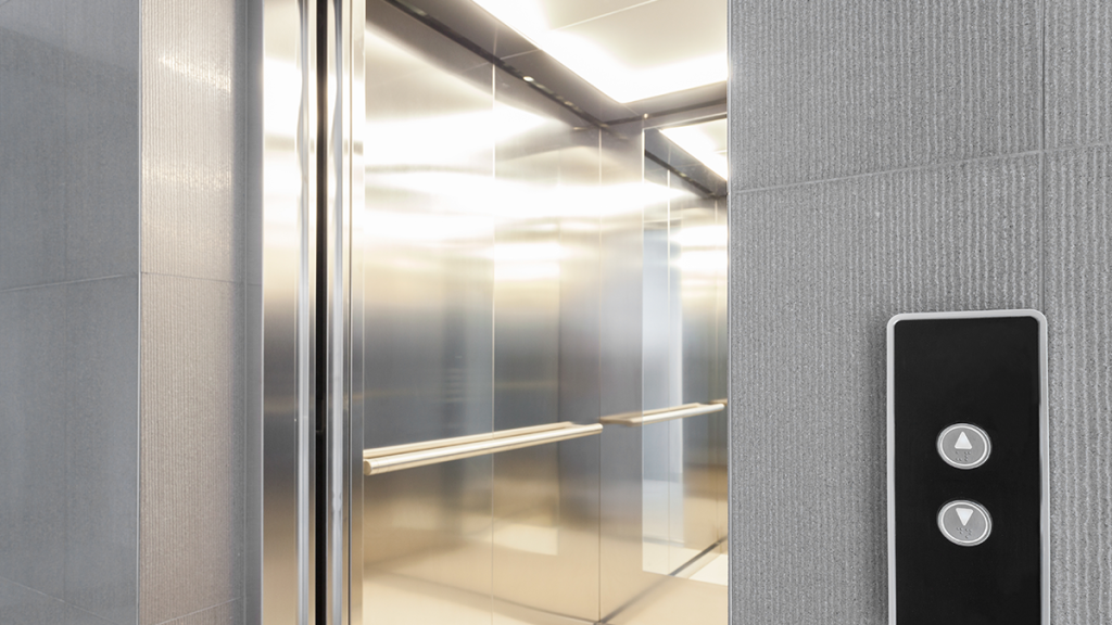 A modern elevator cab interior with polished stainless steel wall panels, a horizontal handrails system, and bright ceiling lighting next to an exterior call button panel.