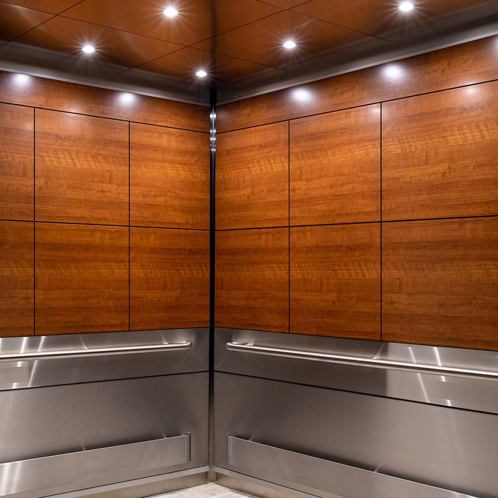 Modern elevator interior featuring SnapCab Apex II design with warm Amber Cherry wood laminate wall panels, stainless steel handrails, and a reflective ceiling with downlighting.